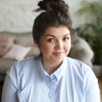 People, youth, joy and happiness concept. Indoor shot of charming chubby young Caucasian woman with hair bun and round cheeks posing in living room, looking at camera with cute joyful smile