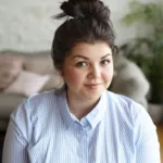 People, youth, joy and happiness concept. Indoor shot of charming chubby young Caucasian woman with hair bun and round cheeks posing in living room, looking at camera with cute joyful smile